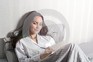 Satisfied and happy smiling girl lying on the bed in the room and writes a journal of your dreams, plans, goals, experiences, idea