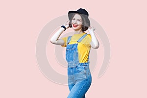 Satisfied hipster woman in overalls, T-shirt, keeps hands on her black hat, smiling to camera.