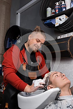 Satisfied man caucasian professional hairdresser washes the hair and head of a client of a young handsome guy with foam before a