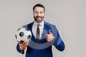 Satisfied man holding soccer ball on his hand with smiling positive expression, showing thumb up.