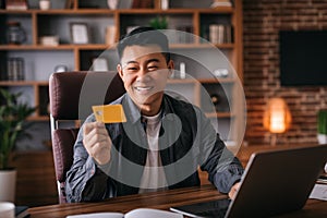 Satisfied middle aged chinese man at table with laptop use credit card in home office interior