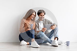 Satisfied millennial caucasian family sitting on floor planning new interior in tablet in empty room