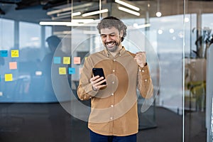 Satisfied millennial male in sandy shirt making victory gesture while looking at screen of mobile phone with happy