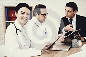 Satisfied nurse typing on tablet in medical office. Blurred doctor and patient in background.