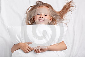 Satisfied playful girl with charming smile, embraces pillow, lies on white pillow, has good rest, enjoys awakening, poses in bed.