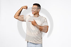 Satisfied, proud assertive young brazilian man with strong muscles, tense raised arm, smiling and pointing biceps