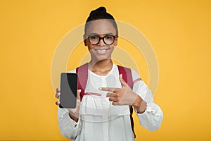 Satisfied smart adolescent black lady pupil in glasses with backpack shows finger at smartphone with blank screen