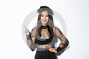 Satisfied smiling asian woman in evil witch or banshee costume celebrating halloween, looking pleased and pointing