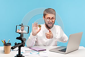 Satisfied smiling man blogger holding golden bitcoin and showing thumbs up posing at smartphone camera, talking about way to make
