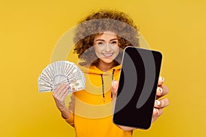 Satisfied smiling woman holding fan of dollar banknotes and showing big smart phone empty display.