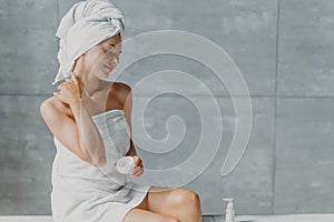 Satisfied spa woman sits in profile applies body cream for perfect skin, enjoys pampering and beauty treatment poses wrapped in