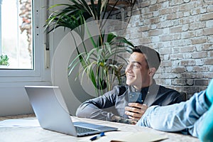 Satisfied teenager happy talking on phone at work with laptop in office, raises hands and puts feet up on table, relaxing after