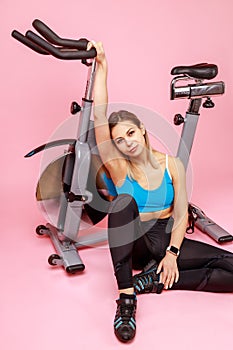 Satisfied tired woman sitting on floor near exercise bike, looking at camera, finishing her workout.