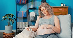 Satisfied woman, dressed in comfortable home clothes, lies on sofa leaning against armrest, stroking pregnant
