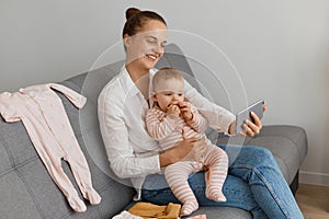 Satisfied woman wearing white shirt and jeans sitting on sofa with her baby daughter, holding cell phone in hands, having online