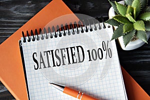 SATISFIED 100%- words in white notebook on dark wooden background with cactus and pen