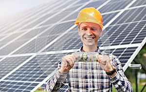 Satisfied worker with hundred dollar bill in hand against the background of solar batteries