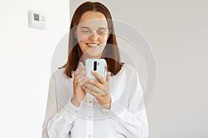 Satisfied young adult female wearing white casual style shirt, posing in light room at home, holding cell phone, looking at gadget