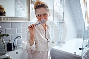 Satisfied young caucasian blonde female thinking with smile while brushing her teeth in the bathroom