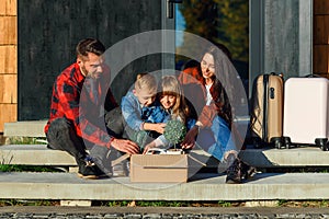 Satisfied young parents with their happy children sitting on new house`s stairs and get from the carton box green flower