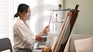 Satisfied young woman painting with watercolor on canvas in bright living room. Art, creative hobby and leisure activity