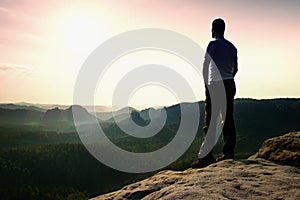 Satisfy tall hiker in grey shirt and dark trousers. Sprtsman on the peak of sharp rock edge watching down to landscape.