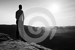 Satisfy tall hiker in grey shirt and dark trousers. Sprtsman on the peak of sharp rock edge watching down to landscape.