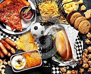 Saturated fats. Football time. TV remote control and snacks - chips, popcorn, cookies, cheese, sauce, fries, burger, nuts