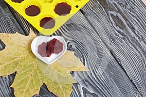 Saucer with cherry marmalade. A silicone mold with marmalade is visible. Dried maple leaves all around. On pine planks painted