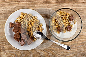 Saucer with porous chocolate and granola, bowl with muesli, yogurt, chocolate, spoon on table. Top view