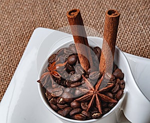 A saucer with a white cup on the table. The cup contains coffee beans, two cinnamon sticks and two star anise.View from above.