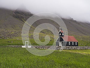 Saurbaejarkirkja black wooden red roof church on green grass field, steep hills in fog, Raudisandur, iceland west fjords