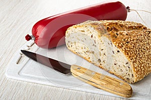 Sausage in polyethylene pack, knife, piece of bread on cutting board on wooden table