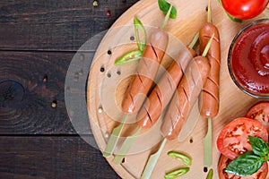 Sausages on skewers with ketchup and fresh tomatoes, served on a round wooden tray on a dark wooden table. Top view.