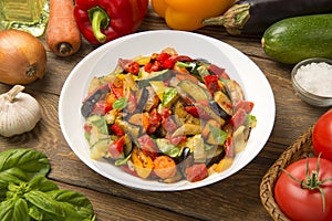 Sautéed eggplant zucchini carrots and tomatoes in a salad bowl on a rustic wooden table with ingredients.