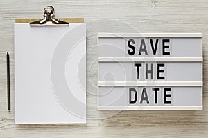 `Save the date` words on a lightbox, clipboard with blank sheet of paper on a white wooden surface, top view. Overhead, from abo