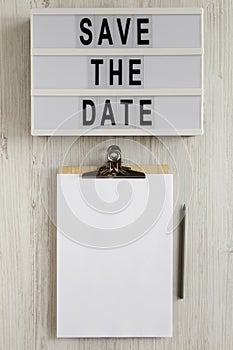 `Save the date` words on a modern board, clipboard with blank sheet of paper on a white wooden background, top view. Overhead,