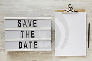`Save the date` words on a modern board, clipboard with blank sheet of paper on a white wooden surface, top view. Overhead, from