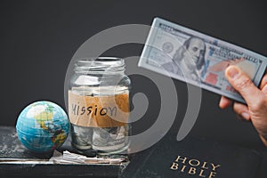 Saving jars full of money and globe with Holy Bible for mission, Mission christian idea. Hand holding dollar with bible on wooden