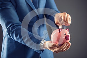 Saving money. Businesman holding pink piggy and putting coin into piggy bank