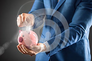 Saving money. Businesman holding pink piggy and putting coin into piggy bank