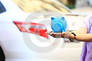 Saving money and loans for car concept, Young woman holding blue piggy with standing at the car parking lot background, Auto