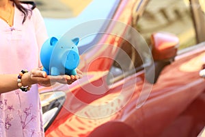 Saving money and loans for car concept, Young woman holding blue piggy with standing at the car parking lot background, Auto