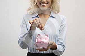 Savings and investment concept. Businesswoman putting money in pink piggy bank, light grey studio background, crop