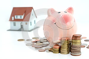 Savings for real estate project with small model house and piggy bank climbing on piles of coins