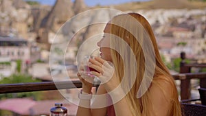 Savor the ambiance of Cappadocia as a young woman enjoys a hot drink on a terrace amidst unique cave houses. The cafe