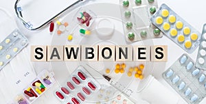 SAWBONES word on wooden blocks on a desk. Medical concept with pills, vitamins, stethoscope and syringe on the background