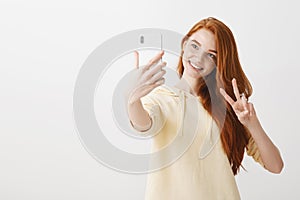 Say hi to technology. Studio portrait of beautiful caucasian girl with ginger hair taking selfie on smartphone, showing