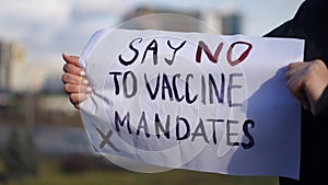 Say no to vaccine mandates placard in female Caucasian hands with blurred urban city at background. Unrecognizable
