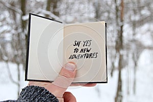 Say yes to new adventures on the background of the winter forest. Hand holding a book with the inscription.